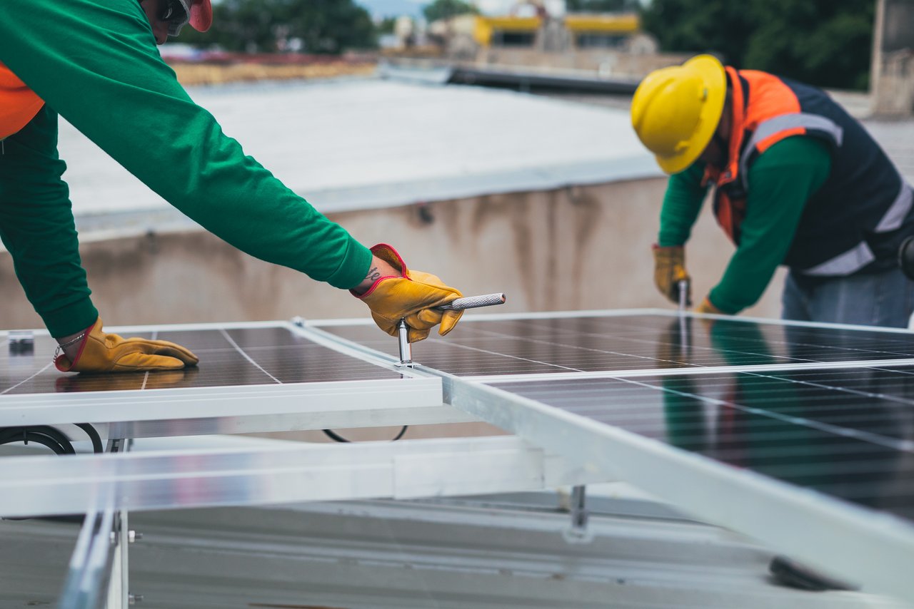 Das Bild zeigt zwei Handwerker in Schutzkleidung, die auf einem Dach PV-Module montieren.